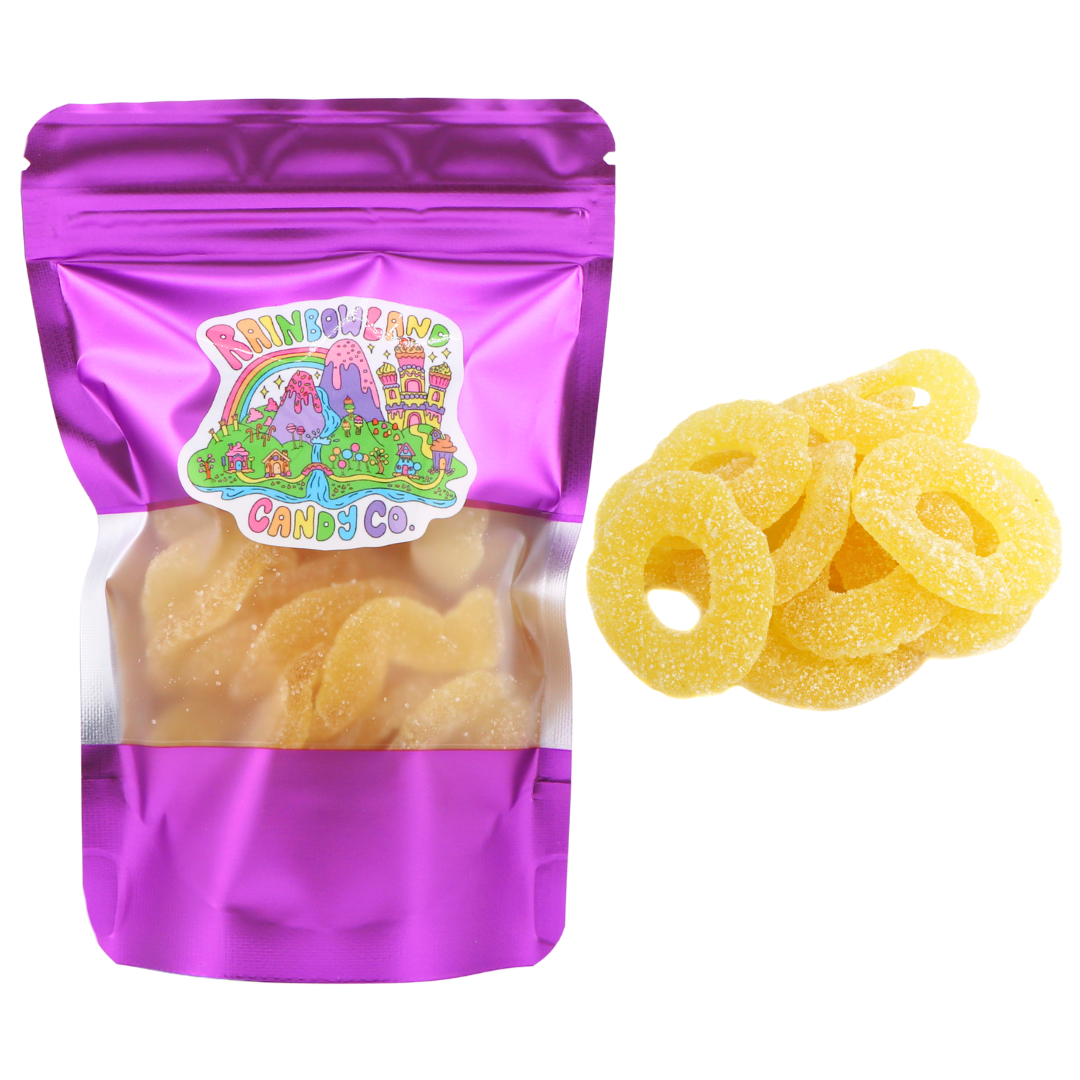 RainbowLand Candy Co. Pineapple Rings in a 4 oz purple metallic resealable stand-up pouch with a clear window, shown beside a loose pile of bright yellow, sugar-coated pineapple-flavored gummy rings.