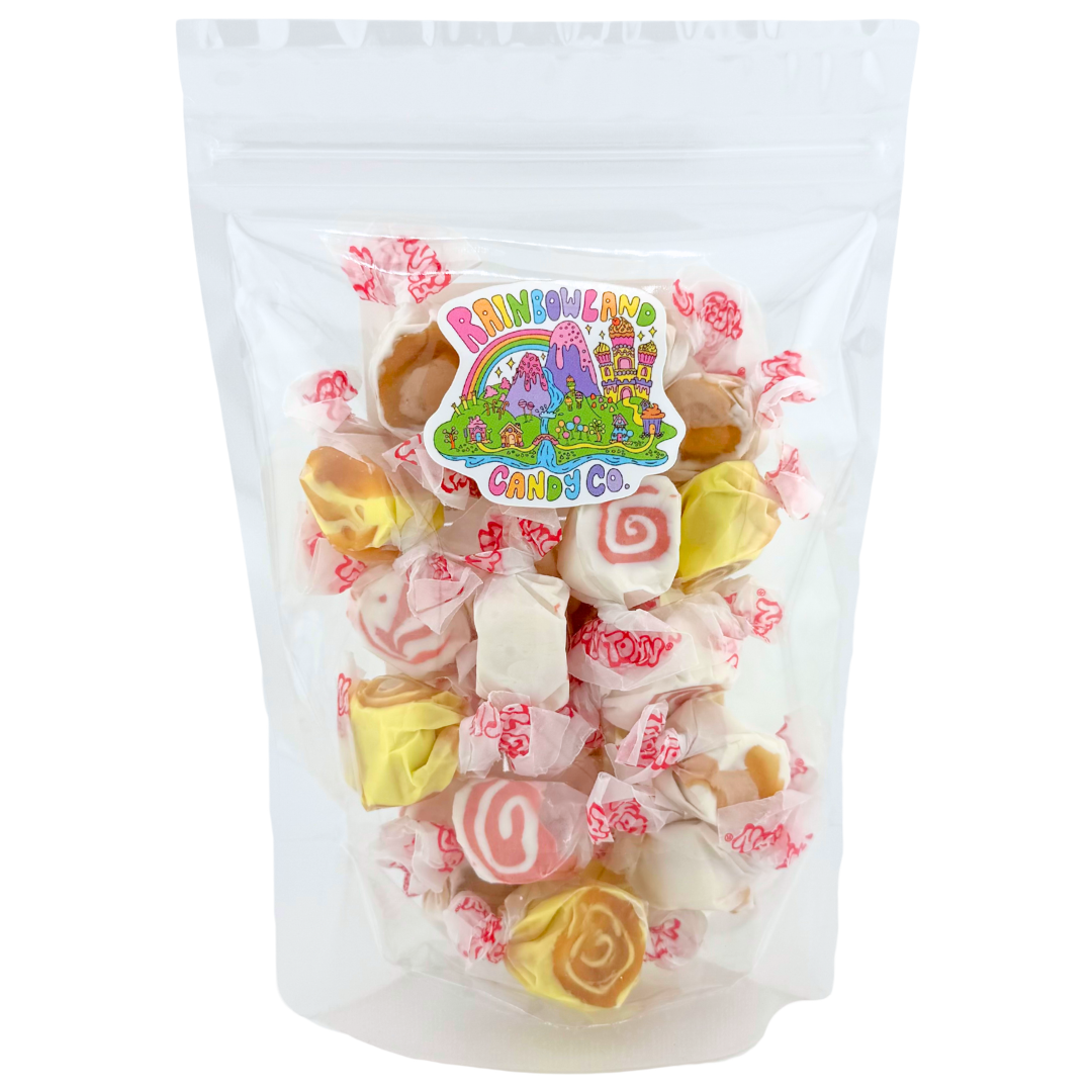 Clear resealable stand-up pouch of RainbowLand Candy Co. Piece of Cake Taffy Trio Mix, filled with individually wrapped Taffy Town salt water taffy in pineapple upside-down cake, red velvet cake, and German chocolate cake flavors, featuring yellow taffy with caramel swirls, white taffy with red spirals, and cream taffy with golden-brown centers, topped with the colorful RainbowLand candy co logo sticker.