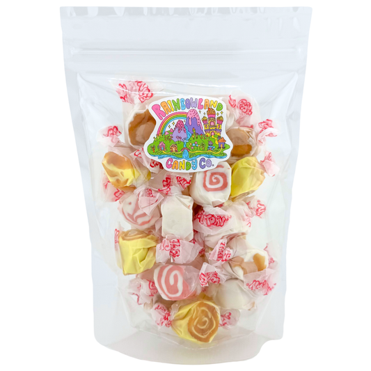 Clear resealable stand-up pouch of RainbowLand Candy Co. Piece of Cake Taffy Trio Mix, filled with individually wrapped Taffy Town salt water taffy in pineapple upside-down cake, red velvet cake, and German chocolate cake flavors, featuring yellow taffy with caramel swirls, white taffy with red spirals, and cream taffy with golden-brown centers, topped with the colorful RainbowLand candy co logo sticker.